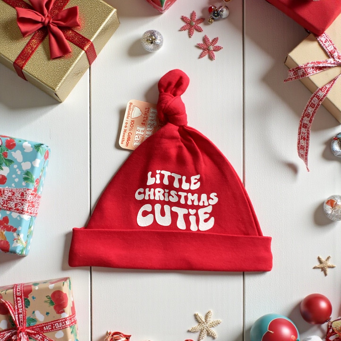Red baby hat with 'Little Christmas Cutie' text on a festive background with presents and ornaments.