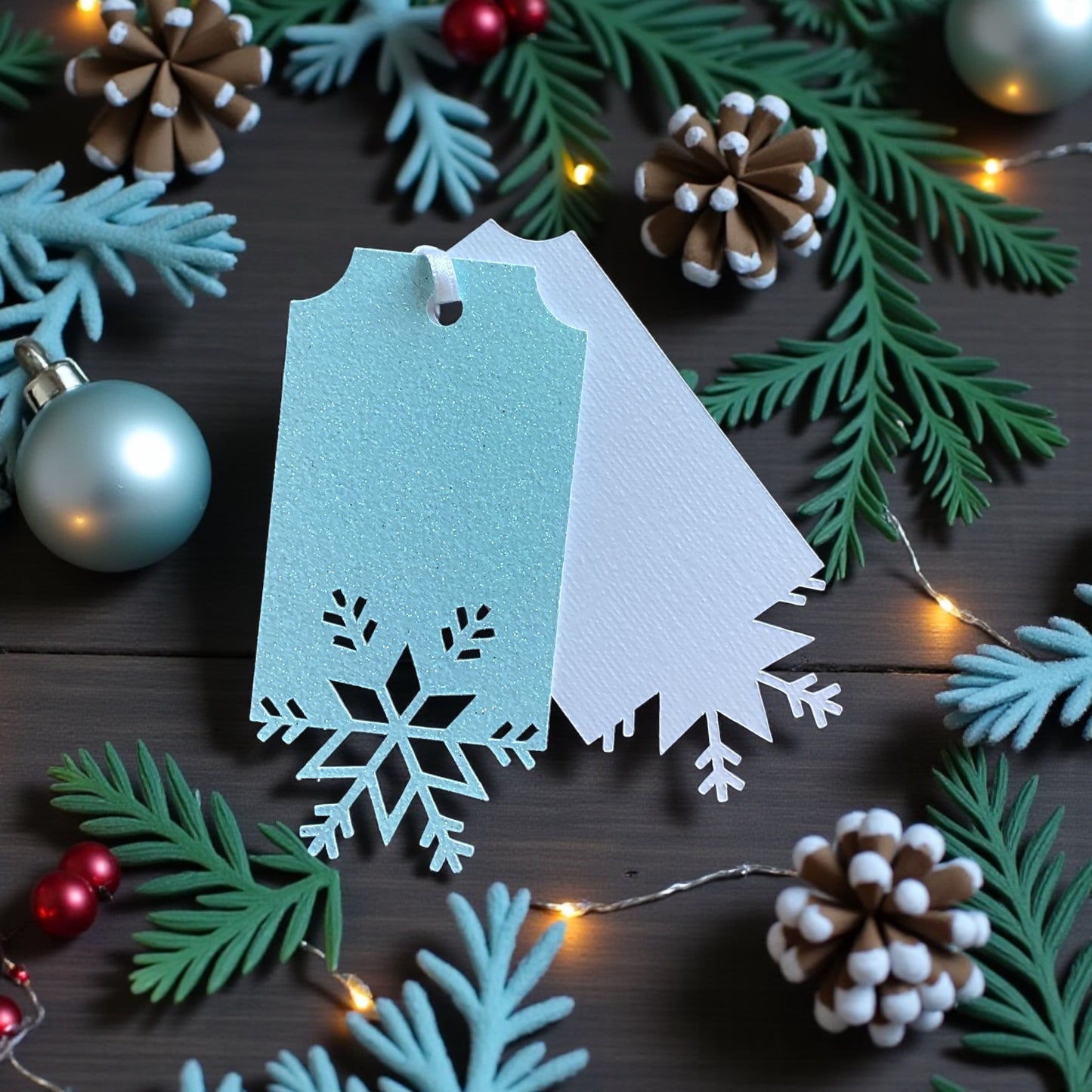 "Close-up of ice blue glitter snowflake gift tag showing perforated snowflake detail and plain white reverse, styled with Christmas baubles and pine branches"