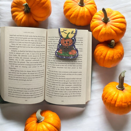 Open book with a decorative pumpkin bookmark surrounded by small pumpkins on a white surface