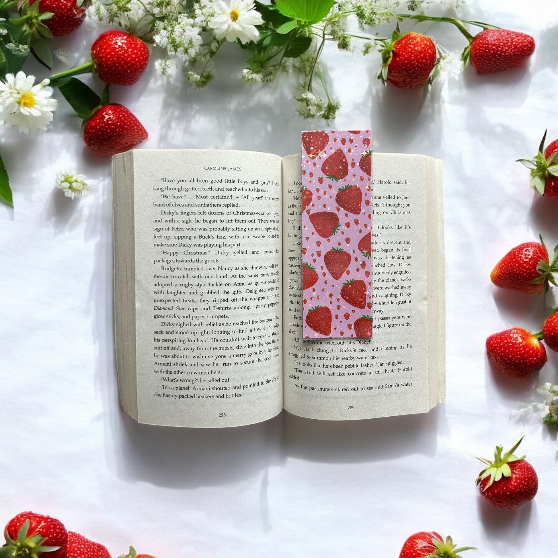 Open book with a strawberry-themed bookmark surrounded by strawberries and flowers on a white surface