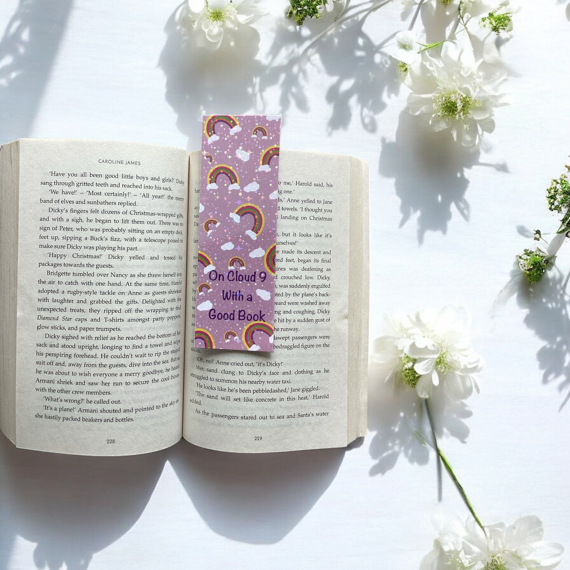 Open book with a colorful bookmark titled 'On Cloud 9 With A Good Book' surrounded by white flowers on a light surface.