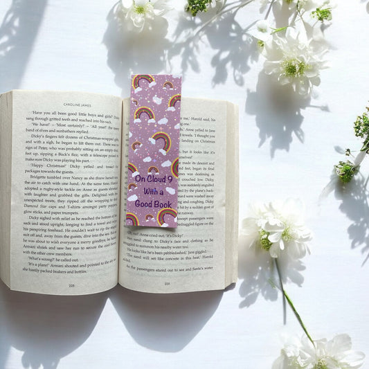 Open book with a colorful bookmark titled 'On Cloud 9 With A Good Book' surrounded by white flowers on a light surface.