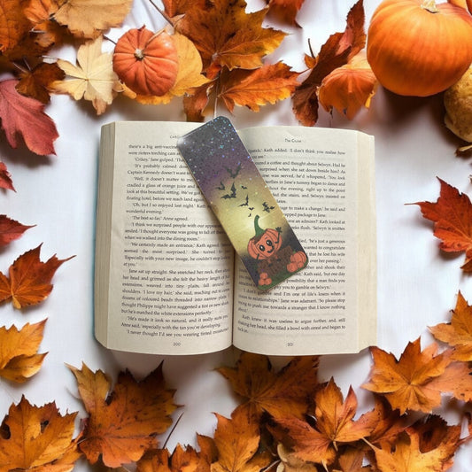 Open book with a pumpkin-themed bookmark surrounded by autumn leaves and pumpkins on a white background