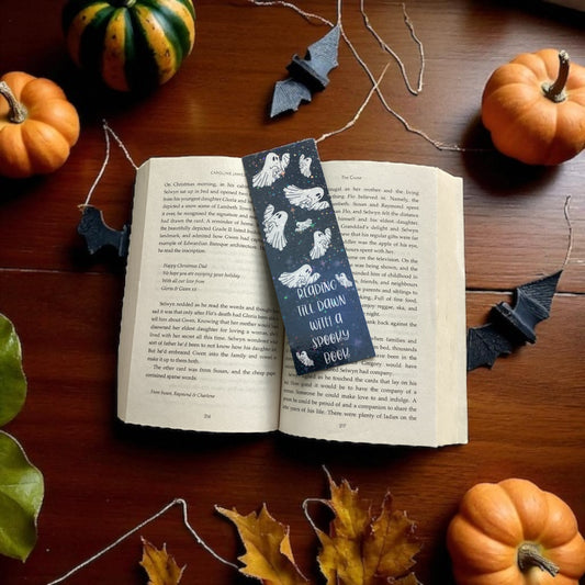 Open book with a bookmark featuring ghosts on a wooden surface with pumpkins and leaves.