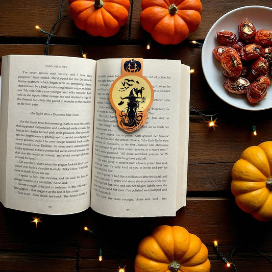 An open book on a dark wooden table with a Halloween-themed decorative bookmark featuring a witch riding a broomstick inside a pumpkin. The scene is styled with small orange pumpkins, a plate of chocolate-covered dates, and warm string lights.