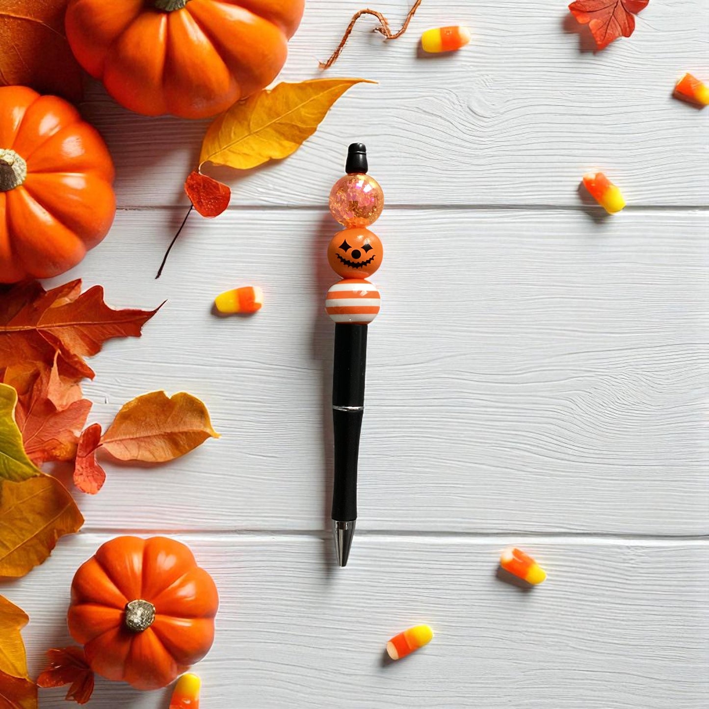 A Halloween-themed pen with orange and white beads featuring a jack-o'-lantern and candy corn design, styled on a white wood background with pumpkins, autumn leaves, and candy corn.