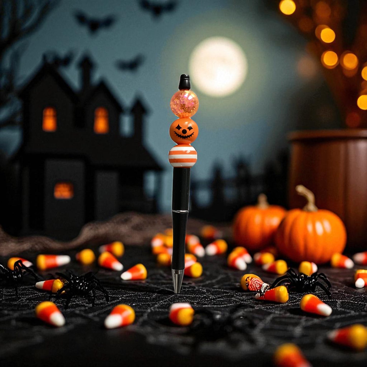 A Halloween-themed pen standing upright on a black surface, decorated with a jack-o'-lantern topper, candy corn, and a pink glitter ball. The background shows a spooky haunted house, pumpkins, cobwebs, and candy under a full moon.