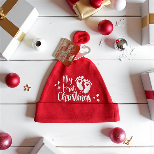 Red baby Santa hat with 'My First Christmas' text on a white wooden surface with Christmas decorations.