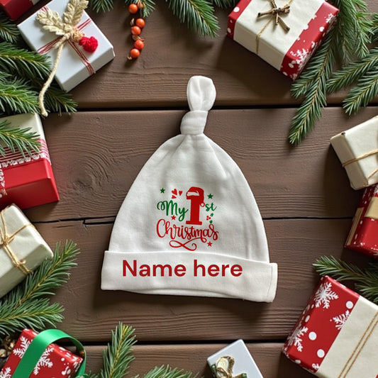 Baby hat with 'My 1st Christmas' text on a wooden table with gift boxes and pine branches.