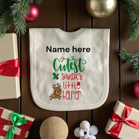 Baby bib with Christmas-themed text and graphics on a wooden surface with festive decorations.