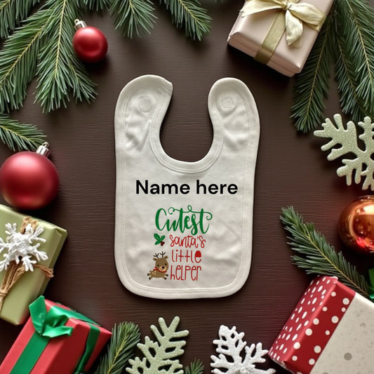 Baby bib with 'Cutest Santa's Little Helper' text on a brown background with Christmas decorations.