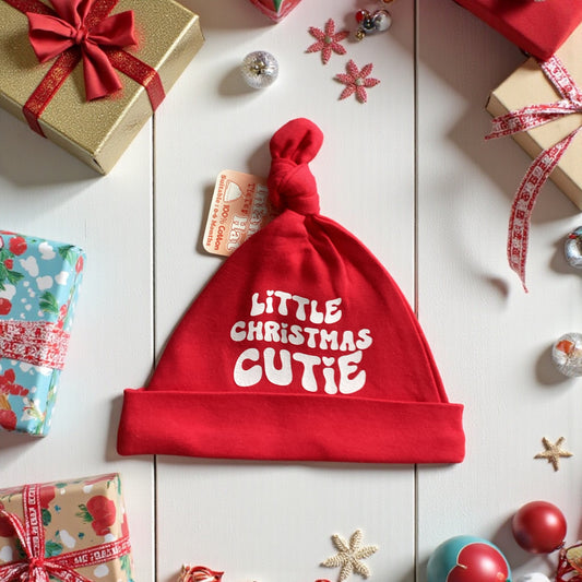 Red baby hat with 'Little Christmas Cutie' text on a festive background with presents and ornaments.