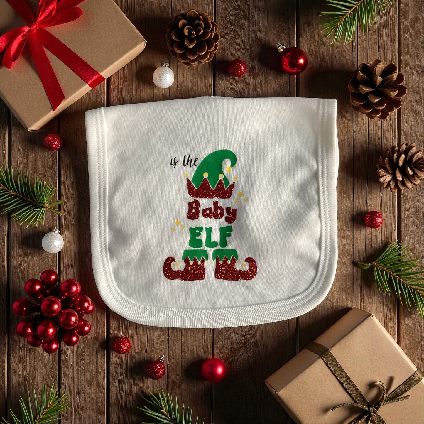 Baby bib with 'Is the Baby Elf' design on a wooden surface with Christmas decorations.