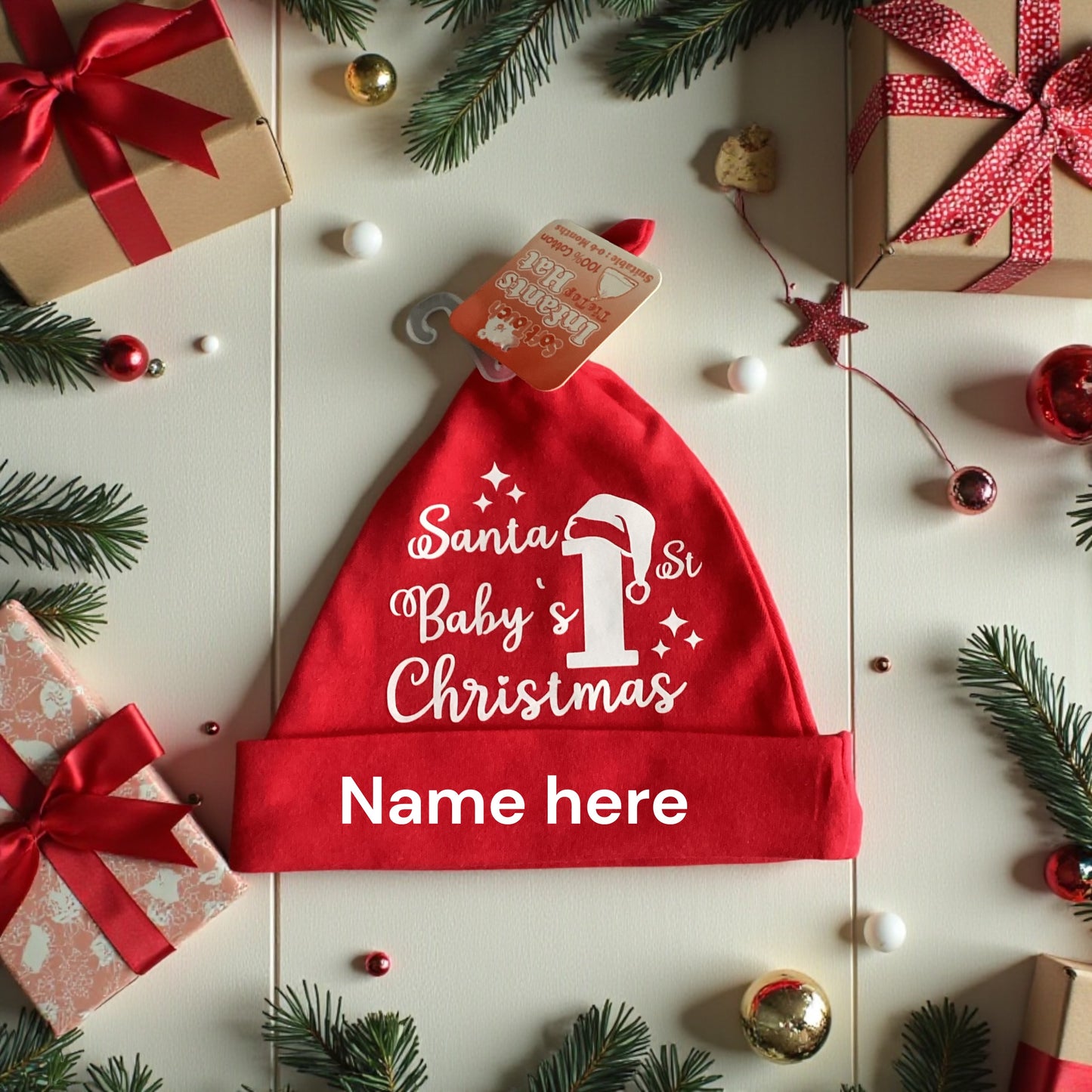 Red personalised baby knotty hat with Santa Baby's 1st Christmas design and name placeholder, surrounded by festive gifts and pine branches