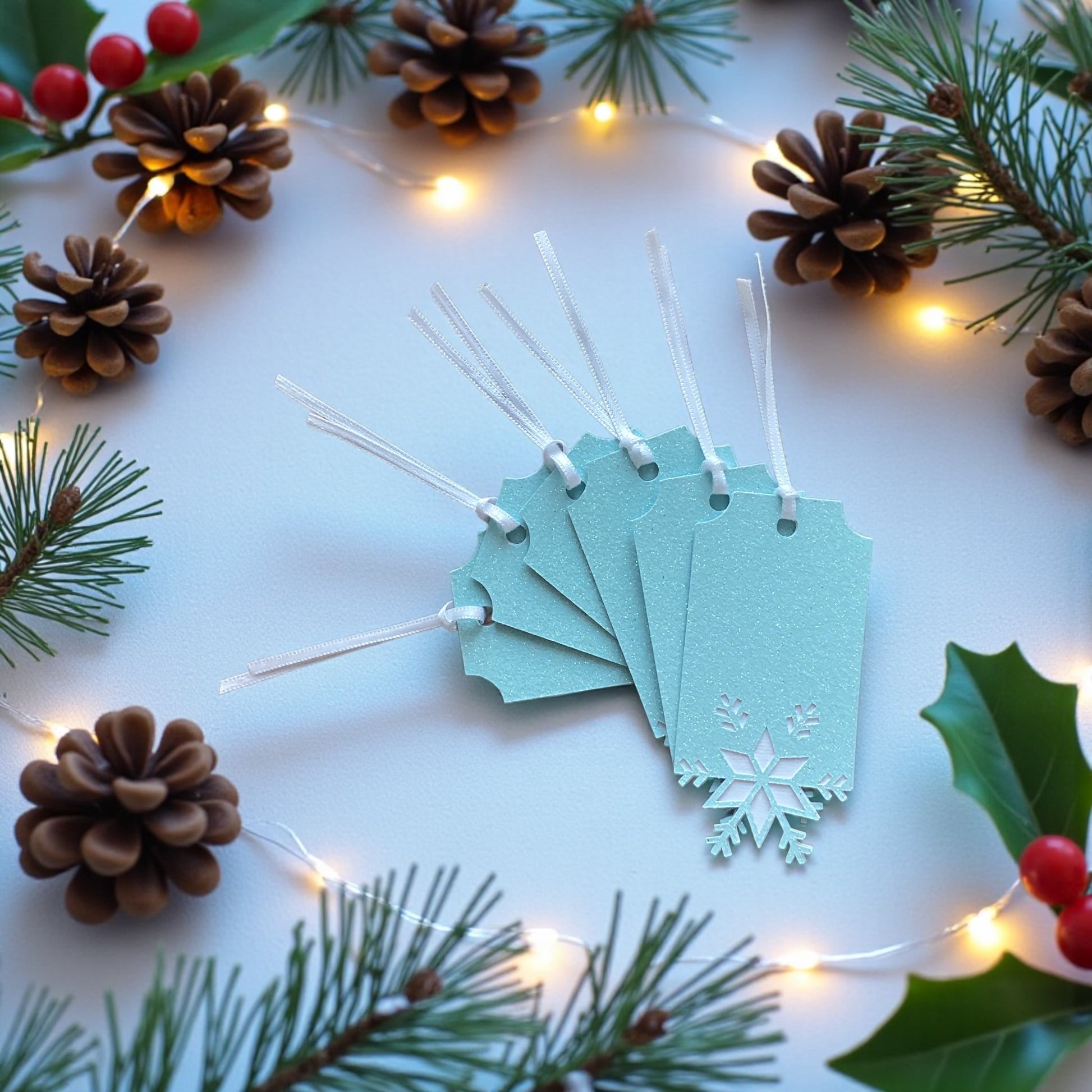 "Set of six ice blue glitter snowflake gift tags with white ribbon fanned out on a white surface surrounded by pine cones, holly and fairy lights"