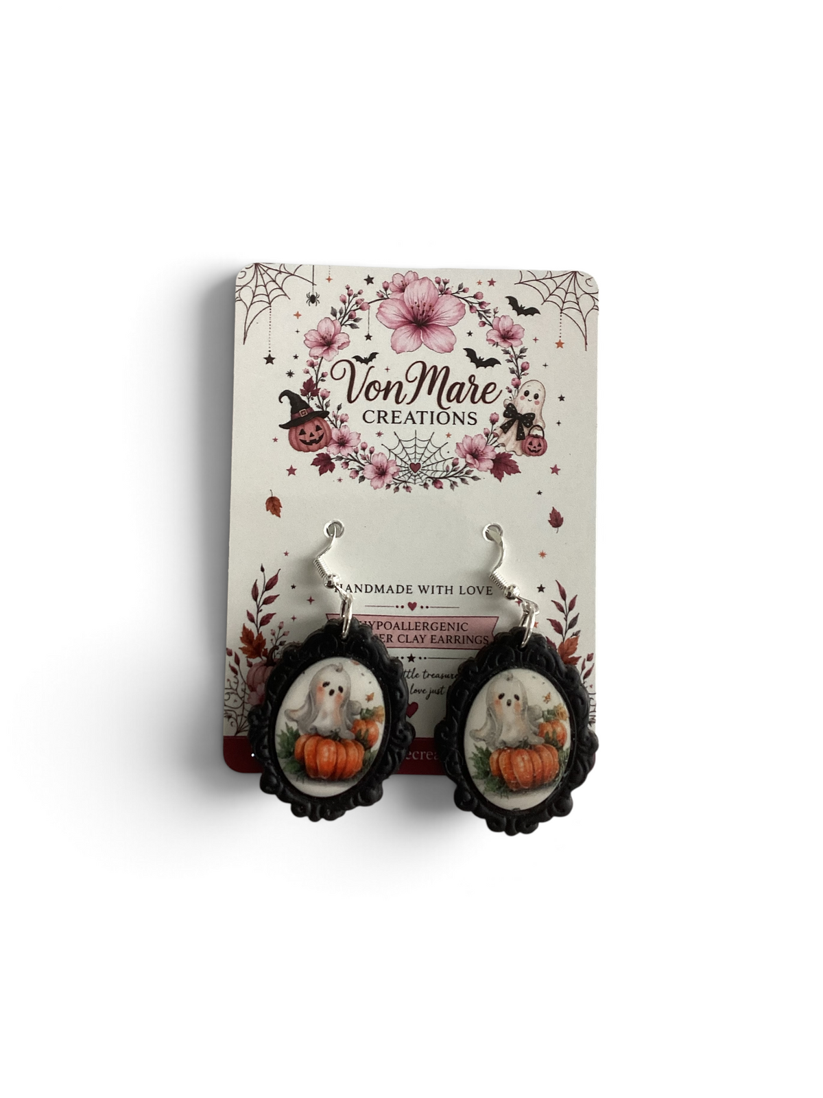 A pair of Halloween-themed dangle earrings displayed on a branded card. The earrings have black scalloped frames and silver hooks, featuring oval charms with a white ghost sitting on an orange pumpkin surrounded by autumn leaves.